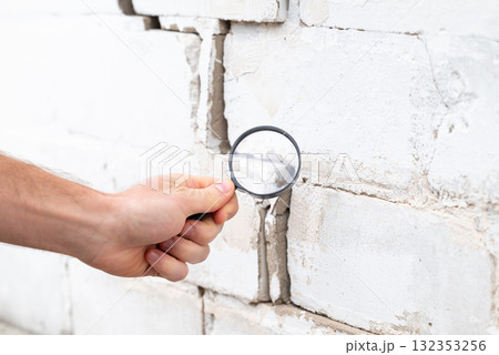 Inspecting building with a crack. Big crack in brick wall, man hands looking at it with magnifying glass Inspecting building with a crack. Big crack in brick wall, man hands looking at it with magnifying glass 132353256