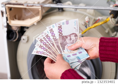 Counting hryvnia cash in front of a disassembled washing machine during repair work. A person counts out cash in front of a washing machine that's currently being repaired, service expenses concept Counting hryvnia cash in front of a disassembled washing machine during repair work. A person counts out cash in front of a washing machine that's currently being repaired, service expenses concept 132353401
