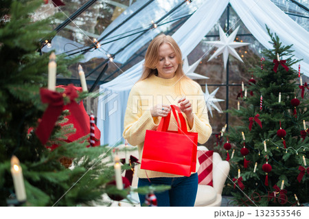 Surprised blonde woman looking into red shopping bags near Christmas tree Surprised blonde woman looking into red shopping bags near Christmas tree 132353546