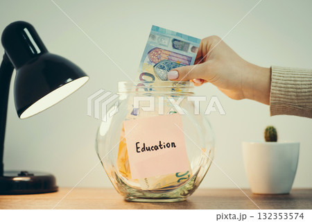 Glass jar with money standing on the table, female hand putting 20 euro banknote inside, lamp and kaktus nearby. Toned close up photo with concepts of saving funds for education Glass jar with money standing on the table, female hand putting 20 euro banknote inside, lamp and kaktus nearby. Toned close up photo with concepts of saving funds for education 132353574