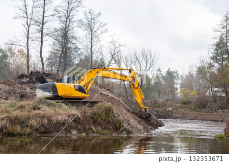 Big industrial excavator digging pond in the countryside. Yellow excavator shoveling dirt from the bottom of an artificial pond 132353671