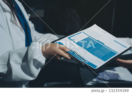 Two doctors and a female nurse meet at a table in the hospital, collaborating on medical tasks using laptops and computers Two doctors and a female nurse meet at a table in the hospital, collaborating on medical tasks using laptops and computers 132354169