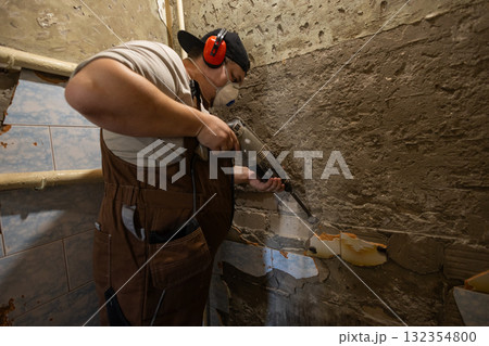 A man wearing a respirator and earmuffs uses a powerful hammer drill to remove old plaster and cement from a wall during a home renovation. Removing cement from walls with a hammer drill. 132354800