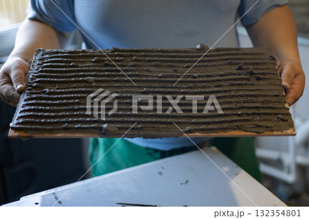 A worker holds a ceramic tile with a fresh layer of ribbed, wet cement adhesive on the back, preparing for installation during a home renovation. The technician holds the tile with the adhesive. A worker holds a ceramic tile with a fresh layer of ribbed, wet cement adhesive on the back, preparing for installation during a home renovation. The technician holds the tile with the adhesive. 132354801