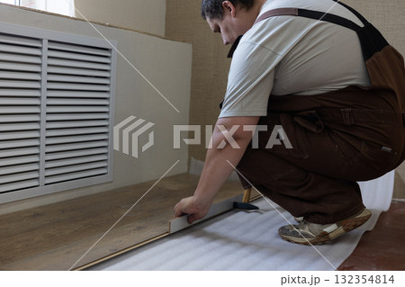 A man in overalls lays laminate flooring over underlayment, installing new flooring during a residential renovation. The man demonstrates his professional flooring installation skills. 132354814
