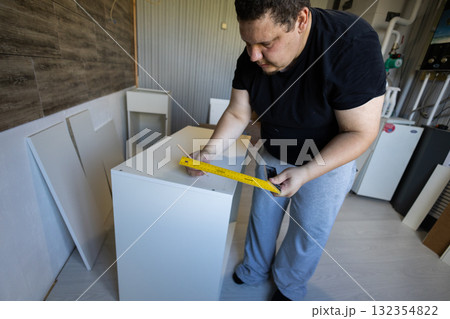 Man measuring and marking kitchen cabinet components with a ruler and pencil during a home renovation, assembling modern white cabinets with careful diy craftsmanship and precision. 132354822