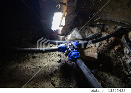 Blue plastic adapter connecting black water pipes for plumbing system repair in a dark, dusty basement, illuminated by a work light, showing maintenance work on an underground utility. 132354824
