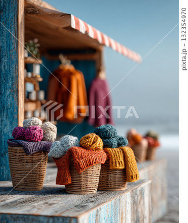 A collection of colorful knitted balls displayed in baskets on a wooden surface, with blurred clothing items and beach scenery in the background 132354970