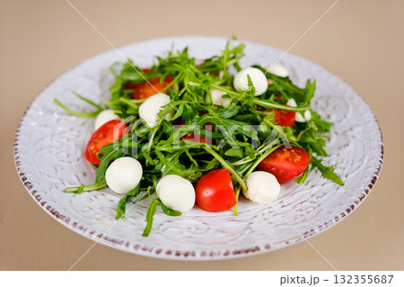 Fresh green salad made with mozzarella cheese, cherry tomatoes, arugula and olive oil on white stone table background. Healthy food and diet concept. Flat lay, copy space. Fresh green salad made with mozzarella cheese, cherry tomatoes, arugula and olive oil on white stone table background. Healthy food and diet concept. Flat lay, copy space. 132355687