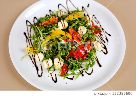 Caprese salad with arugula, mozzarella, cherry tomatoes, fresh basil and balsamic glaze on a white plate on a gray concrete background. Top view, copy space Caprese salad with arugula, mozzarella, cherry tomatoes, fresh basil and balsamic glaze on a white plate on a gray concrete background. Top view, copy space 132355690