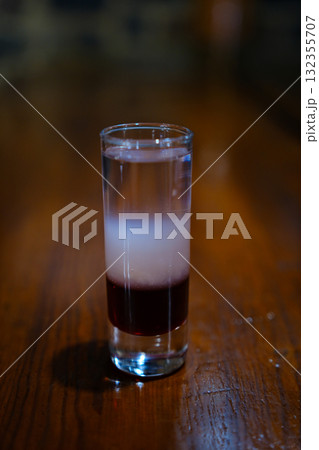 Colorful layered cocktail shot glass with orange, dark, and light layers surrounded by melting ice cubes on dark glossy bar surface, perfect for nightlife and party themes. 132355707