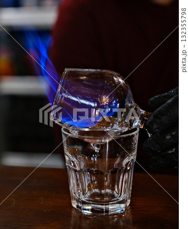 Bartender makes hot cocktail. crystal goblet with delicious red alcoholic cocktail that two men bartenders impressively sets to fire at dark bar Bartender makes hot cocktail. crystal goblet with delicious red alcoholic cocktail that two men bartenders impressively sets to fire at dark bar 132355798