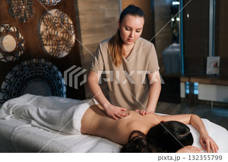 Professional massage therapist performing deep massaging technique on female client's back in serene cozy spa environment, promoting relaxation and healing. Concept of rest with spa treatment. Professional massage therapist performing deep massaging technique on female client's back in serene cozy spa environment, promoting relaxation and healing. Concept of rest with spa treatment. 132355799