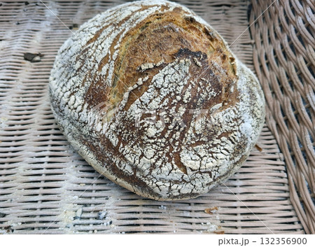 A round loaf of homemade farm bread sits on a woven basket. The crust is golden brown and has a rustic texture, showcasing the art of traditional baking at its finest. A round loaf of homemade farm bread sits on a woven basket. The crust is golden brown and has a rustic texture, showcasing the art of traditional baking at its finest. 132356900
