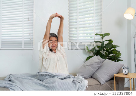 Stretching and Wellness. An elderly woman performing morning stretches on a couch, embracing a healthy lifestyle. Stretching and Wellness. An elderly woman performing morning stretches on a couch, embracing a healthy lifestyle. 132357804