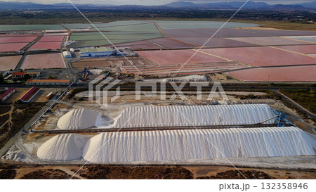 Aerial drone view of colorful salt evaporation ponds and white salt piles near Santa Pola, Spain, with industrial structures and mountains in the distance. 132358946