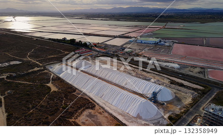 Drone view of large white salt piles and colorful evaporation ponds near Santa Pola, Spain, with mountains and reflections in evening light. 132358949
