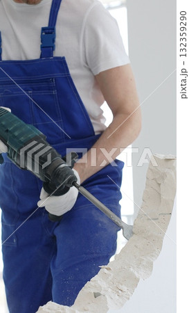 Close up of unknown male construction worker wearing blue overalls is demolishing white wall with rotary hammer drill, generating dust, close up horizontal view. Renovation concept Close up of unknown male construction worker wearing blue overalls is demolishing white wall with rotary hammer drill, generating dust, close up horizontal view. Renovation concept 132359290