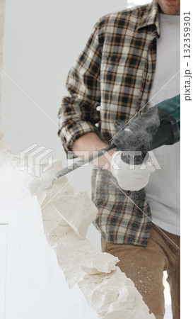 Close up of unrecognizable male construction worker wearing beige checkered shirt and protective gloves, is demolishing white wall with rotary hammer drill, generating dust. Renovation concept 132359301