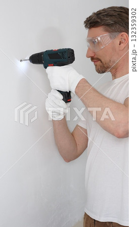 Middle aged man worker wearing white t-shirt and protective gloves, is drilling hole in a wall with cordless drill during home renovation work. Vertical portrait view Middle aged man worker wearing white t-shirt and protective gloves, is drilling hole in a wall with cordless drill during home renovation work. Vertical portrait view 132359302