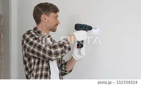 Middle aged man wearing checked shirt and white gloves, is drilling hole in a wall with cordless drill during home renovation work. Portrait view Middle aged man wearing checked shirt and white gloves, is drilling hole in a wall with cordless drill during home renovation work. Portrait view 132359324