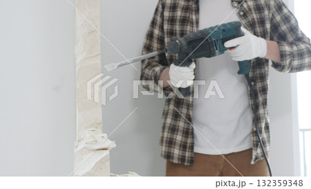Unrecognizable male construction worker wearing beige checkered shirt and protective gloves, is demolishing white wall with rotary hammer drill, generating dust, close up view. Renovation concept 132359348
