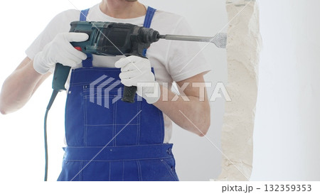 Close up of unrecognizable male construction worker wearing blue overalls and protective white gloves, is demolishing white wall with rotary hammer drill, generating dust, horizontal view. Renovation 132359353