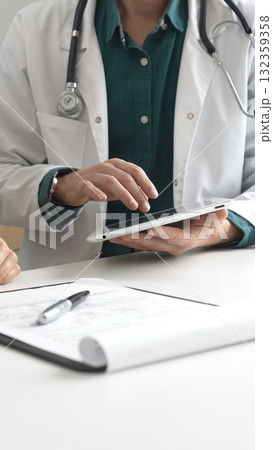 Male doctor in lab coat over a green shirt, and stethoscope gesturing while explaining diagnosis to patient using tablet in modern medical office. Medicine and health care concept 132359358