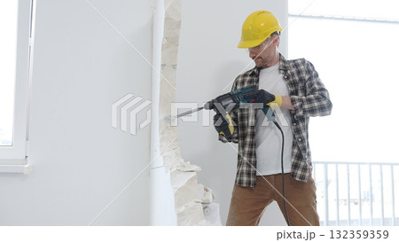 Man construction worker wearing beige checkered shirt , yellow hard hat and protective gloves, is demolishing white wall with rotary hammer drill, generating dust, medium long shot. Renovation concept 132359359