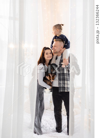 A happy family poses together, with a child on the father's shoulders and a dog in the mother's arms, in a cozy, well-lit room. A happy family poses together, with a child on the father's shoulders and a dog in the mother's arms, in a cozy, well-lit room. 132366244