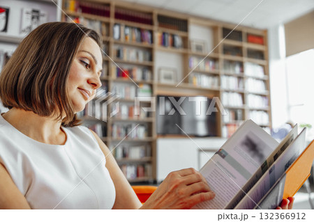 A young Caucasian woman with brown hair reads a book in a modern library. Bookshelves filled with various books are visible in the background. 132366912