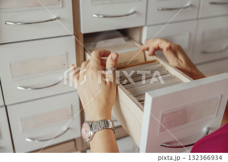 A person with light skin and short brown hair organizes index cards in a wooden drawer. The setting is a modern library or coworking space. A person with light skin and short brown hair organizes index cards in a wooden drawer. The setting is a modern library or coworking space. 132366934