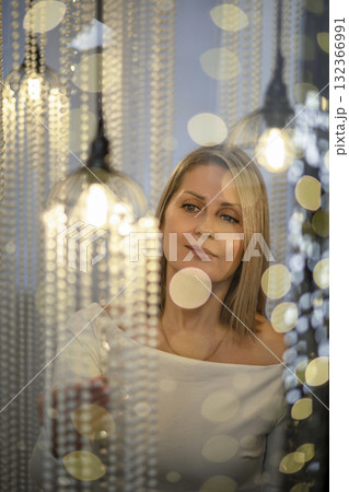 A young Caucasian woman with blonde hair gazes thoughtfully amidst sparkling Christmas lights. The scene conveys a magical holiday atmosphere. A young Caucasian woman with blonde hair gazes thoughtfully amidst sparkling Christmas lights. The scene conveys a magical holiday atmosphere. 132366991