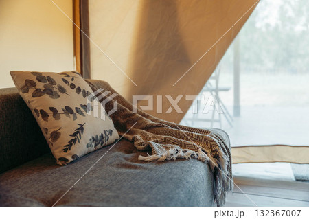 Cozy glamping tent interior with a sofa, decorative pillow, and a soft blanket. Natural light filters through the tent fabric, creating a warm atmosphere. Cozy glamping tent interior with a sofa, decorative pillow, and a soft blanket. Natural light filters through the tent fabric, creating a warm atmosphere. 132367007