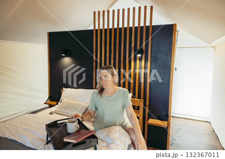 A young Caucasian woman with brown hair sits on a bed in a glamping tent. She holds a cup and has a book on a tray beside her. The interior features wooden accents. A young Caucasian woman with brown hair sits on a bed in a glamping tent. She holds a cup and has a book on a tray beside her. The interior features wooden accents. 132367011