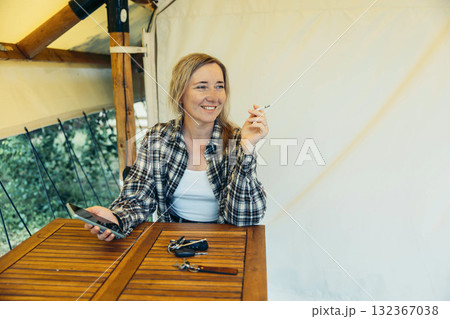 Mindful mornings in nature Peaceful start of the day in tent cabin. woman sitting inside glamping tent holding cigarette and phone on wooden chair at rustic table with casual outfit Mindful mornings in nature Peaceful start of the day in tent cabin. woman sitting inside glamping tent holding cigarette and phone on wooden chair at rustic table with casual outfit 132367038