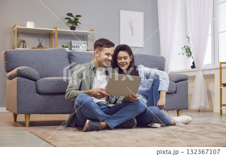 Young happy family couple sitting on floor in modern living room, looking at laptop screen 132367107