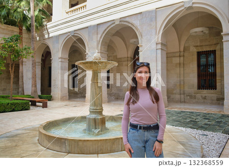 Mediterranean courtyard with fountain, surrounded by classical arches and columns. A stylish person in purple top and jeans explores this serene space Mediterranean courtyard with fountain, surrounded by classical arches and columns. A stylish person in purple top and jeans explores this serene space 132368559