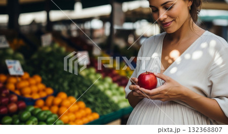 Smiling pregnant woman holding a red apple while shopping for fresh, healthy fruits and vegetables at farmers market. Smiling pregnant woman holding a red apple while shopping for fresh, healthy fruits and vegetables at farmers market. 132368807