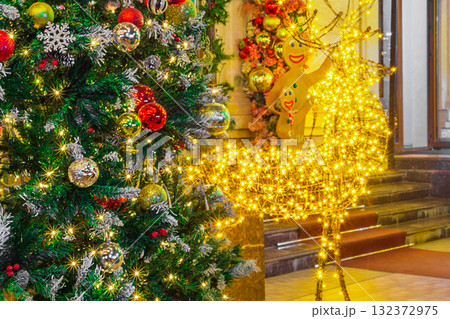 Decorated Christmas or New Year's tree, adorned with Christmas tree ornaments and garlands that glow and sparkle, creating bokeh effect, next to a figure of a deer and two figures of gingerbread men Decorated Christmas or New Year's tree, adorned with Christmas tree ornaments and garlands that glow and sparkle, creating bokeh effect, next to a figure of a deer and two figures of gingerbread men 132372975