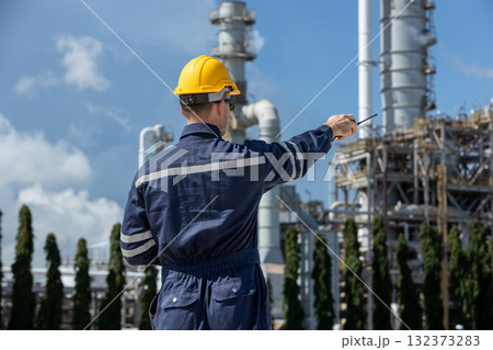 Back view of engineer in safety uniform pointing at refinery plant while using walkie-talkie representing industrial field control, communication, supervision safety management and oil gas operations. 132373283