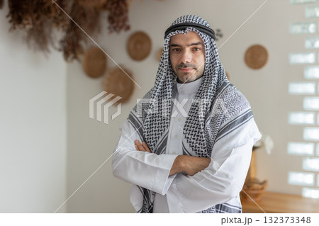 Muslim man or businessman in traditional attire Middle East stands confidently in contemporary indoor space adorned with dried plants and wooden decor. Combination highlights cultural heritage. 132373348