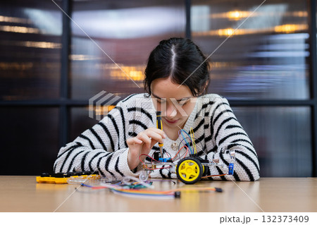Student girl concentrates on assembling robotic car with wires and components at workshop. Engineering classroom with technology and enhancing creativity and learning. STEM education concept. Student girl concentrates on assembling robotic car with wires and components at workshop. Engineering classroom with technology and enhancing creativity and learning. STEM education concept. 132373409