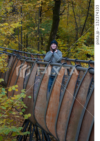 Woman in a black beanie and green quilted jacket standing on the eco trail bridge at VDNH park, surrounded by golden autumn trees 132374103