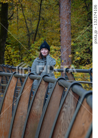Woman in a black beanie and green quilted jacket standing on the eco trail bridge at VDNH park, surrounded by golden autumn trees Woman in a black beanie and green quilted jacket standing on the eco trail bridge at VDNH park, surrounded by golden autumn trees 132374106
