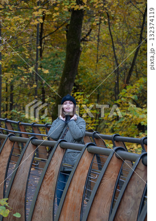 Woman in a black beanie and green quilted jacket standing on the eco trail bridge at VDNH park, surrounded by golden autumn trees 132374119
