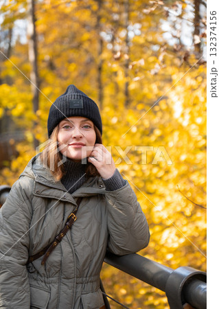 Woman in a green quilted jacket and black knit hat leaning on a wooden bridge surrounded by bright yellow autumn trees 132374156