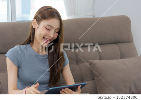 Young happy woman at home enjoys activity on tablet, people find pleasure in digital interaction, relaxing on comfortable sofa, experiencing modern leisure lifestyle 132374800