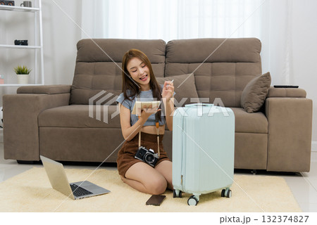 Young happy woman at home plans travel activity, writing trip plans in notebook, enthusiastically preparing for exciting vacation with camera laptop phone and light blue suitcase Young happy woman at home plans travel activity, writing trip plans in notebook, enthusiastically preparing for exciting vacation with camera laptop phone and light blue suitcase 132374827