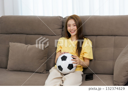 Young happy woman enjoys home activity, people like her watch football, holding soccer ball on comfortable sofa for relaxation indoors Young happy woman enjoys home activity, people like her watch football, holding soccer ball on comfortable sofa for relaxation indoors 132374829